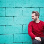 A cheerful man in a red sweater sits against a turquoise wall, enjoying a moment of happiness and laughter.